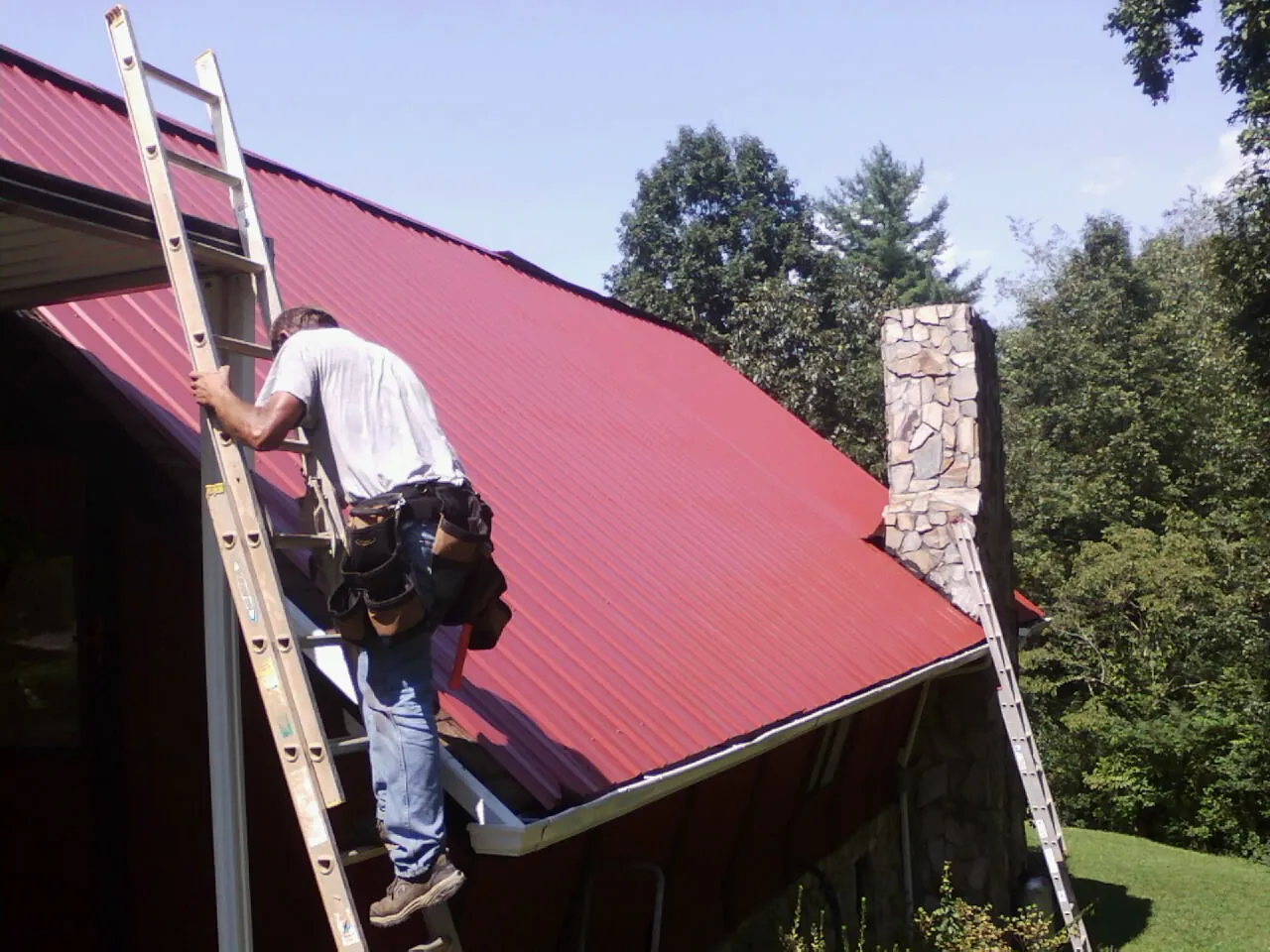 Roofer on a ladder beside a red metal roof and stone chimney — Soffit & Fascia Repair by Roofing Brothers in West Simsbury